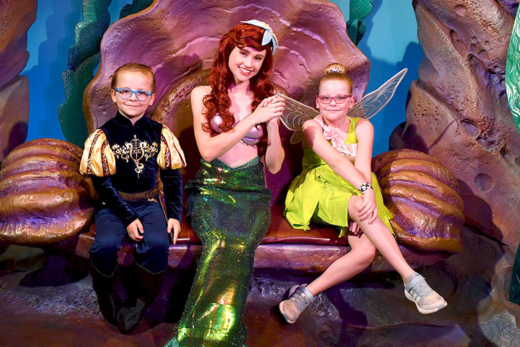 a young boy dressed as a knight and a young girl dressed as Tinker Bell sitting on either side of mermaid Ariel in Magic Kingdom