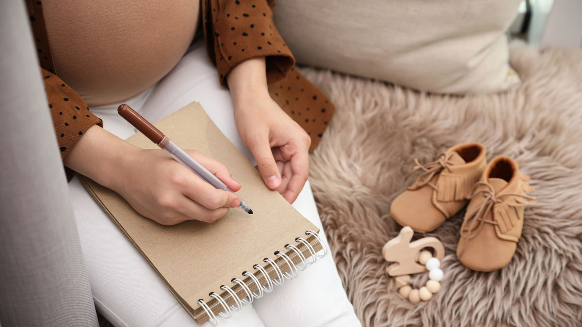 a pregnant woman trying to come up with something to write on her notepad with baby items sitting next to her