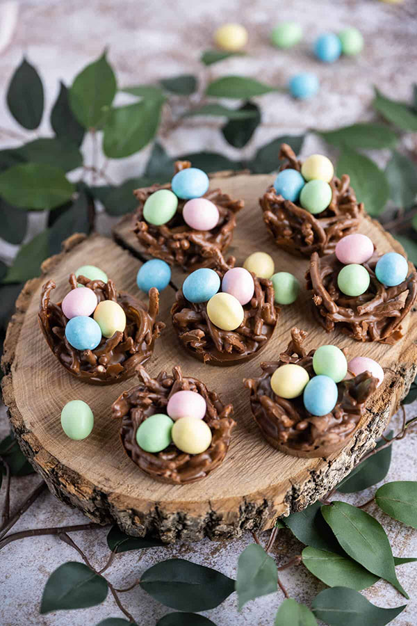 a wood slice platter covered in chocolate cookies resembling birds' nests with mini egg candies on top