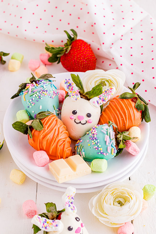chocolate-covered strawberries on a white plate, each decorated for Easter, including a white chocolate bunny face, an orange chocolate made to resemble a carrot, and pastel chocolates