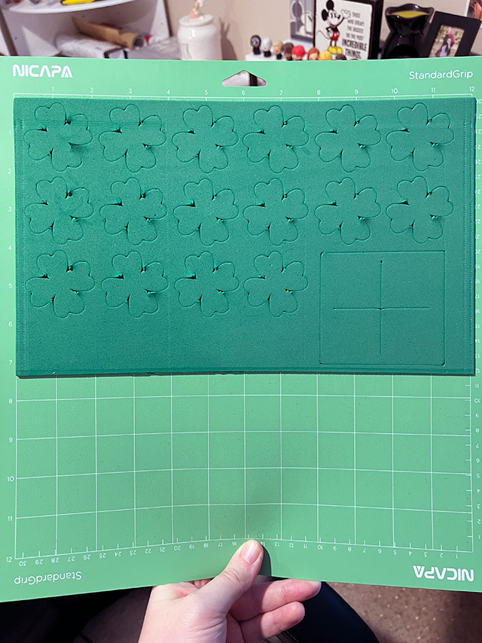 a cutting mat with green craft foam attached that shows shamrocks and a square cut from the foam with a Cricut