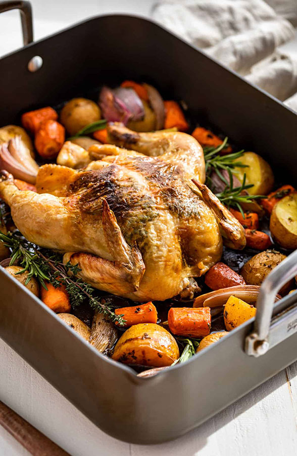 a roasting pan with a cooked chicken and various vegetables surrounding it