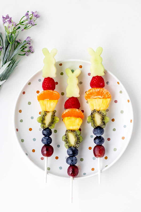 a white polka dot plate with three fruit kabobs side by side, with a piece of honeydew cut into the shape of a bunny at the top of each