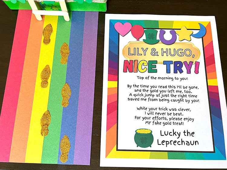 a rainbow road leading to a leprechaun trap with gold-glitter leprechaun footprints leading to it, with a note to the kids lying next to it