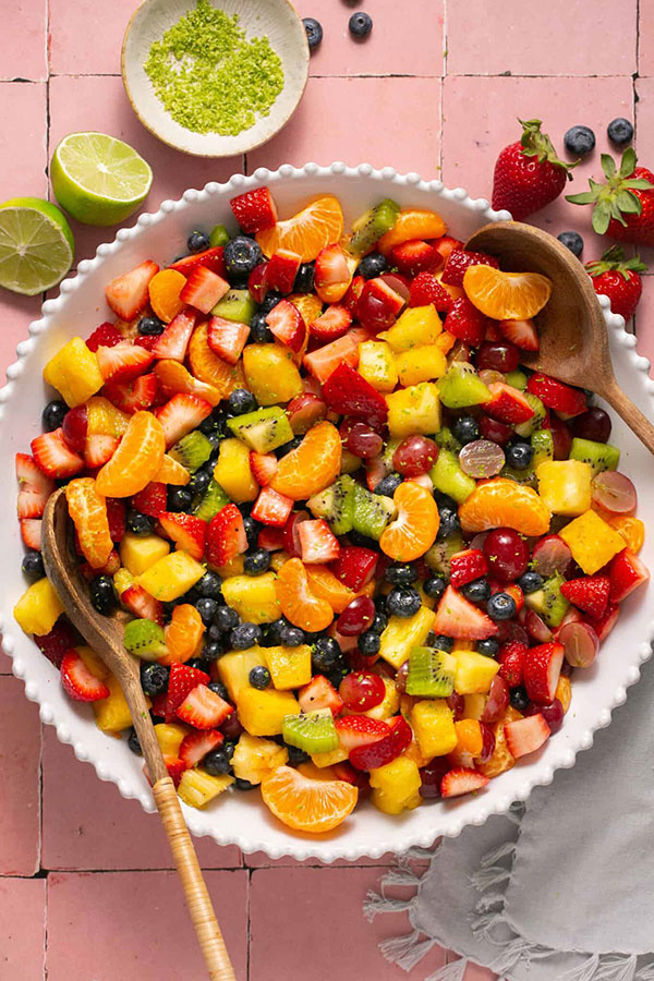 a white bowl full of fruit salad containing strawberries, mandarin oranges, grapes, mango, blueberries, and kiwi