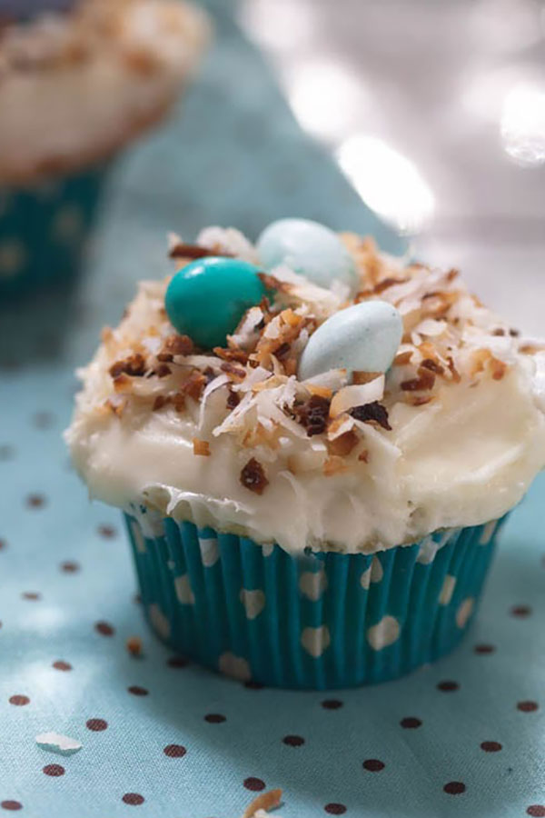 a cupcake in a blue and white polka dot wrapper with white frosting, toasted coconut topping and mini candy eggs on top