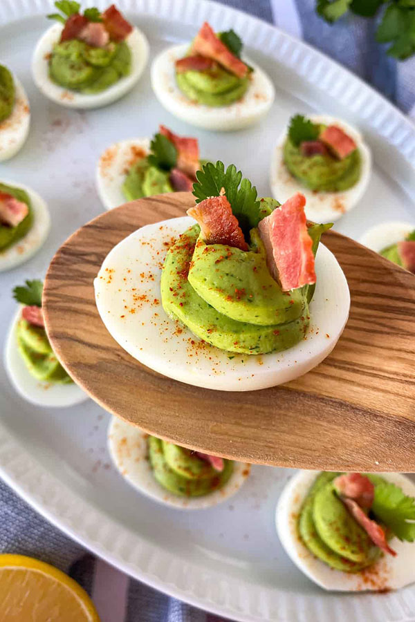 a deviled egg held up on a flat wooden spoon, but the filling is green from avocado and bacon pieces are sticking out of it