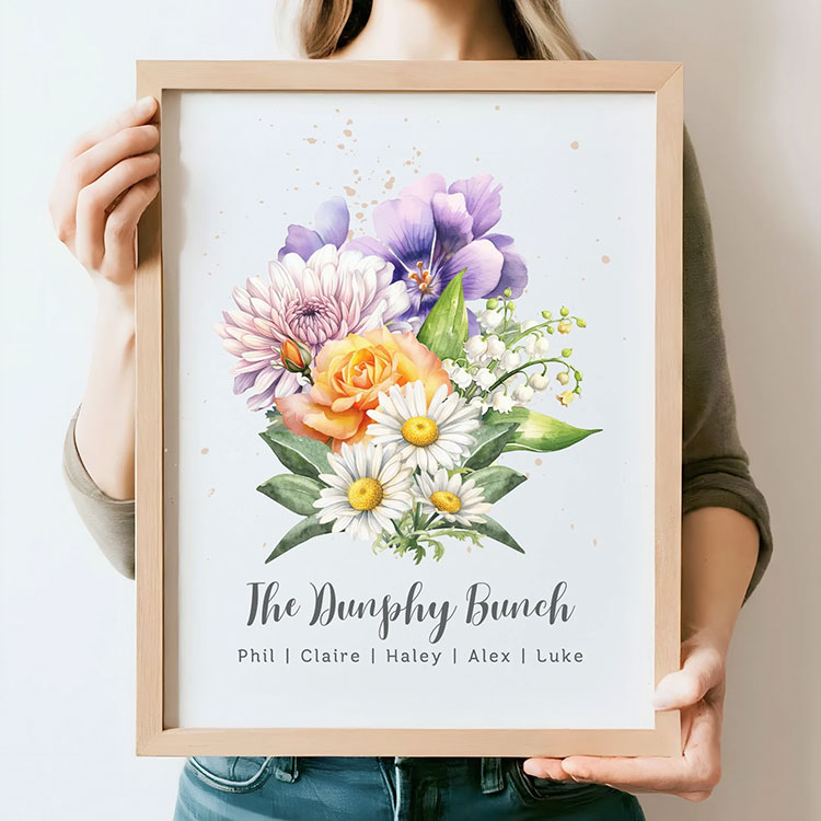 a woman holding a framed watercolor print of various flowers with family names at the bottom