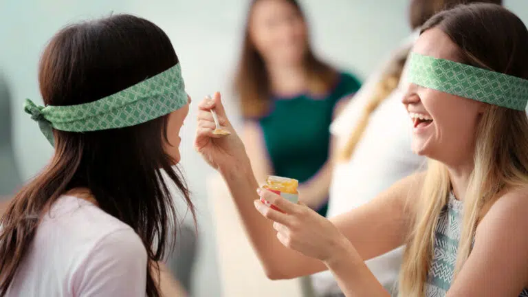 two blindfolded women playing a baby shower game, one trying to feed the other baby food