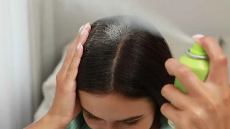 dark-haired woman spraying dry shampoo onto her roots