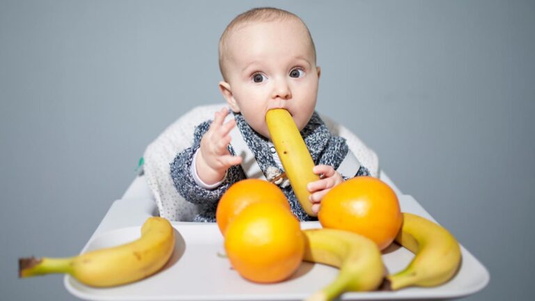 baby sitting in a high chair putting a whole unpeeled banana in its mouth, with the high chair table full of whole bananas and oranges