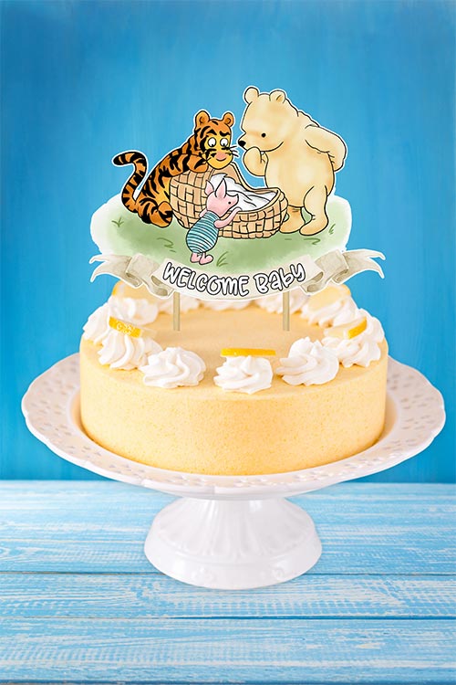 a simple yellow-frosted cake on a white stand with a cardstock topper featuring Pooh, Tigger, and Piglet looking over a baby bassinet with a banner reading "Welcome Baby"