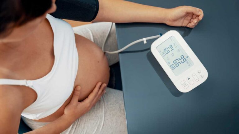 a visibly pregnant woman sitting at a table monitoring her blood pressure with a portable device