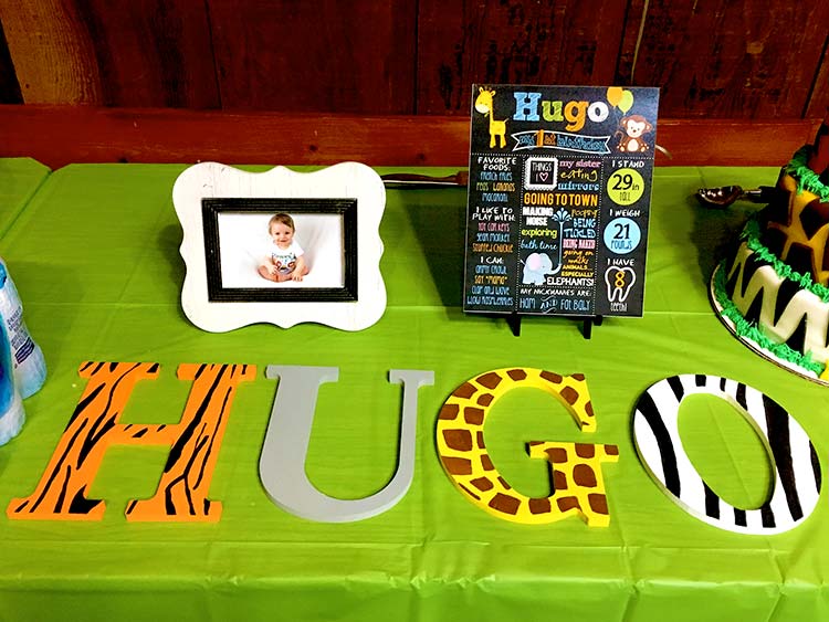 a safari-themed party table with a framed photo, info chalkboard, and wooden letters spelling a name, each letter painted a different animal print