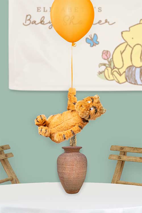 a classic Tigger plush that is attached to a vase so that it appears to be floating up on an orange balloon