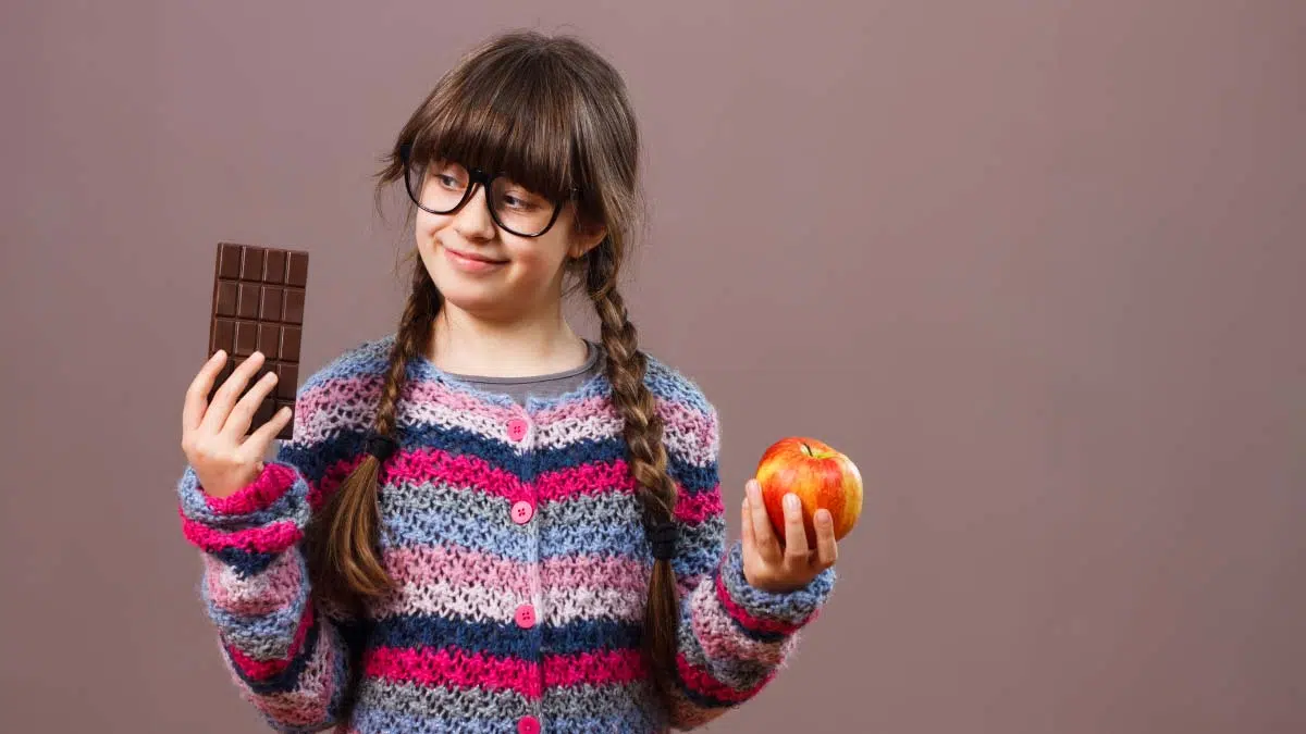 a young girl in braids holding up a bar of chocolate in one hand and an orange in the other