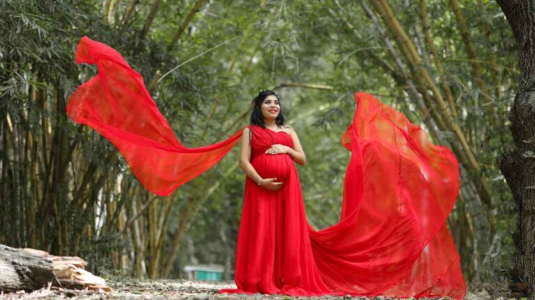 a woman in a vibrant red maternity dress standing in a wooded path with the wind blowing long pieces of red fabric around her