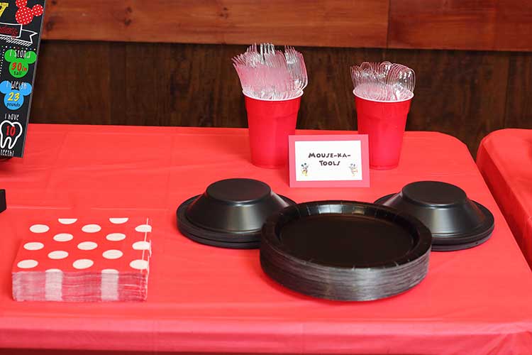 black plates and black bowls arranged to resemble Mickey Mouse's head, red napkins with white polka dots, and cutlery with a card that says "Mouse-ka-tools"