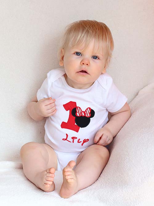 a one-year-old girl wearing a white onesie with a red "1" and a Minnie Mouse head with the name "Lily" embroidered on the body