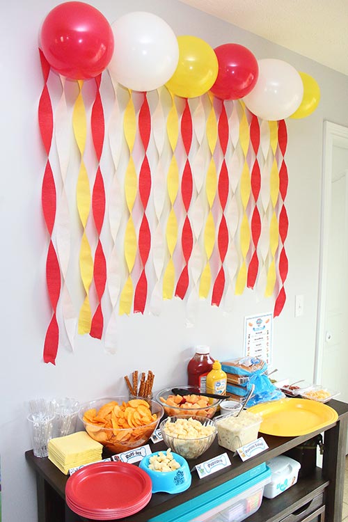 red, white, and yellow balloons hanging high above a kitchen server full of food, with streamers of the same colors dangling below them