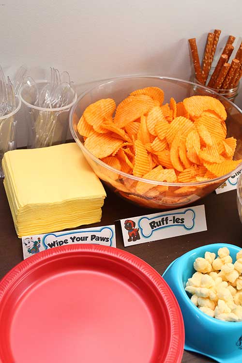 a stack of napkins labeled "Wipe Your Paws", a stack of red paper plates, and a bowl of cheddar Ruffles