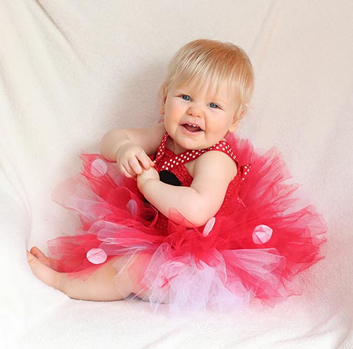 a one-year-old girl sitting in a red, white, and black tutu dress with a Mickey Mouse head silhouette on the chest