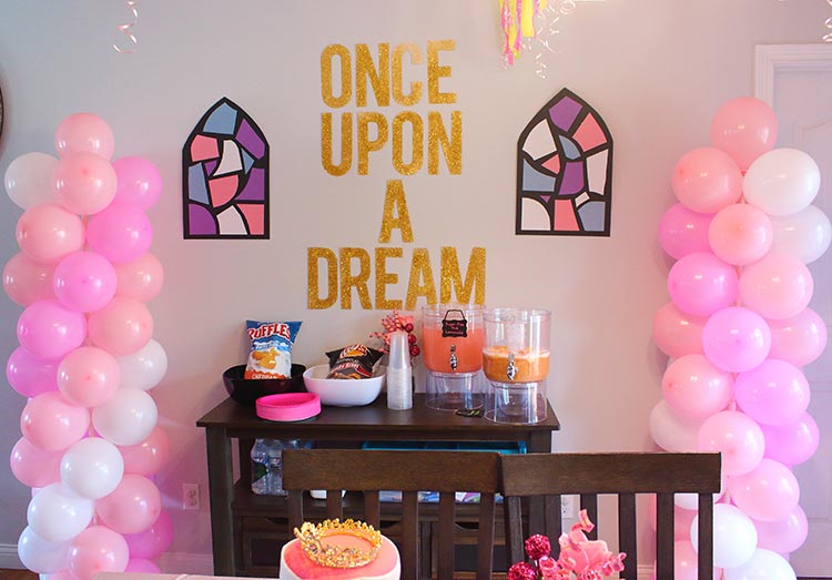 pink and white balloon columns flank a kitchen server and above it are paper stained-glass windows and the words "Once Upon a Dream" in gold glitter