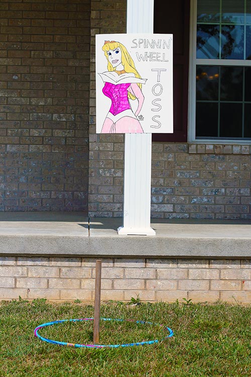 a sign hanging on a porch column featuring a drawing of Princess Aurora and the words "Spinning Wheel Toss" with a stake ringed by a hula hoop on the ground below it
