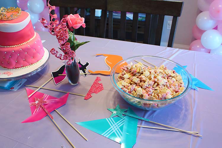 a lavender table with a pink two-tier cake, faux flower arrnagement, photo props from Sleeping Beauty, and pink and white Chex mix
