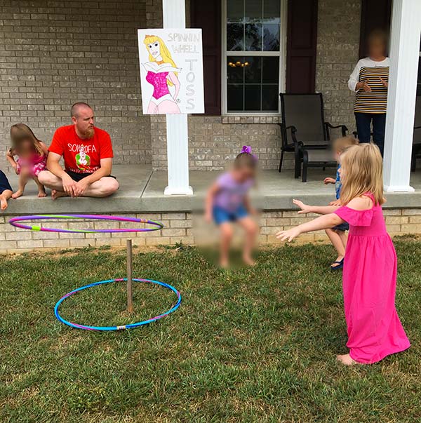 a little girl in a pink dress throwing a hula hoop over a stake in the ground; a sign on the porch behind her features Princess Aurora and says "Spinnin' Wheel Toss"