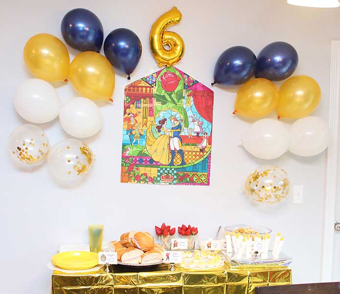 a backdrop at a Beauty and the Beast party featuring navy, gold, and white balloons and a stained-glass window placed over a server filled with themed party foods