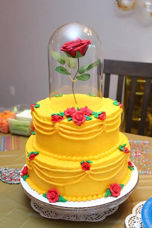 a Beauty and the Beast birthday cake resembling Belle's yellow gown covered in small roses with one large rose on top that appears to be floating covered in a glass cloche