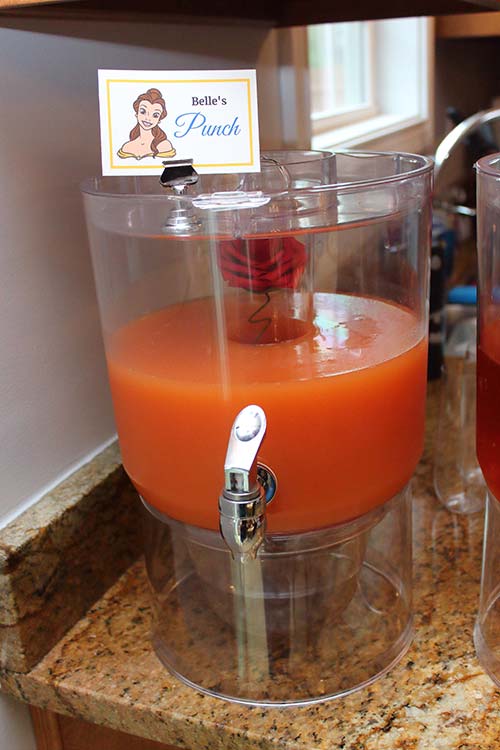 a rose-gold colored party punch labeled "Belle's Punch" with a silk red rose visible in the ice chamber of the drink dispenser