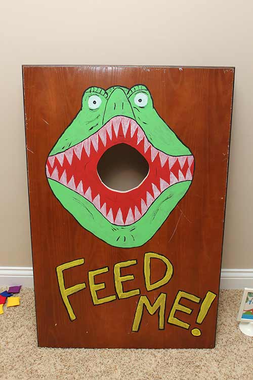 a homemade cornhole-like board with a dinosaur face opening its mouth around the hole with the words "Feed Me!"