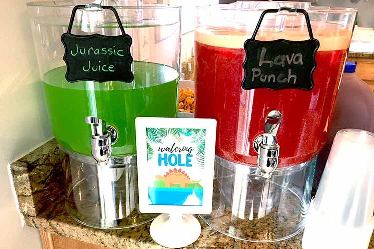 two drink dispensers, one with green liquid labeled "Jurassic Juice" and one with foamy red liquid labeled "Lava Punch" and a framed sign that says "watering hole" with an illustrated stegosaurus