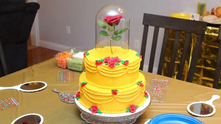 a Beauty and the Beast birthday cake resembling Belle's yellow gown covered in small roses with one large rose on top that appears to be floating covered in a glass cloche