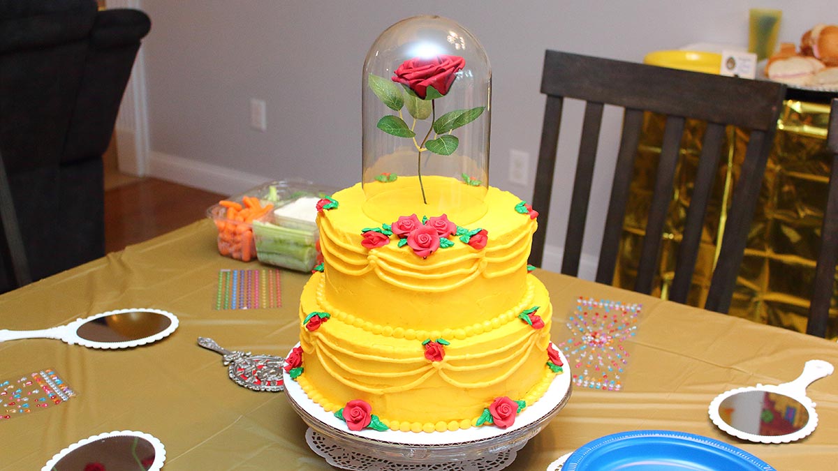 a Beauty and the Beast birthday cake resembling Belle's yellow gown covered in small roses with one large rose on top that appears to be floating covered in a glass cloche