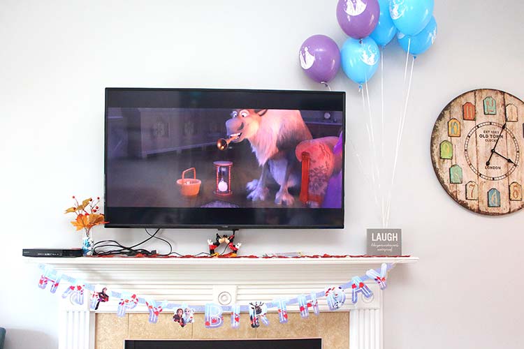 a TV playing Frozen 2 with balloons floating next to it and a Frozen birthday banner hanging under the mantle