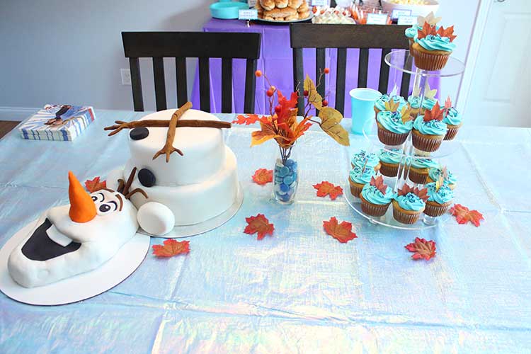 a kitchen table decorated for a Frozen birthday party with an iridescent table cover, Olaf birthday cake, fall leaf decor, and a cupcake stand