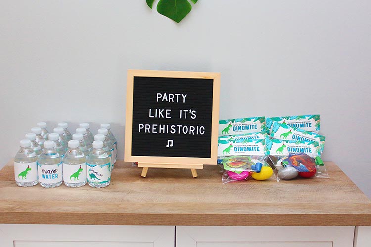 bottles of water with dinosaur labels, a letterboard that says "Party Like It's Prehistoric", and party favor bags filled with silly putty eggs and dinosaur party blowers