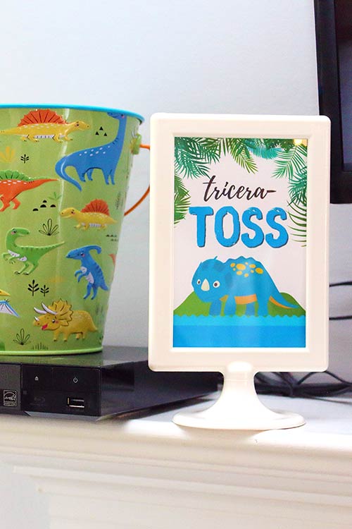 a dinosaur-print metal bucket sitting next to a framed small sign that says "tricera-toss" with an illustrated triceratops