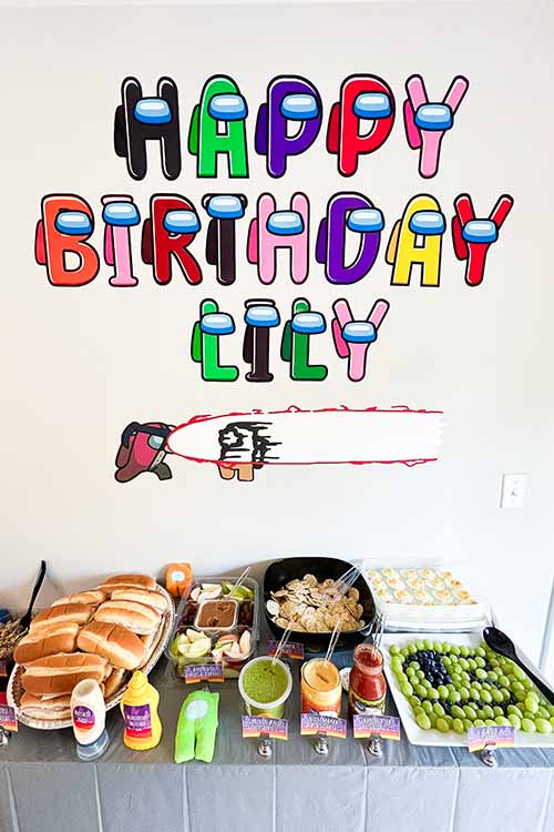 A sign of letters resembling Among Us crewmates says "Happy Birthday Lily" over a cardstock cyborg kill scene. These are all on the wall above a server full of party food.