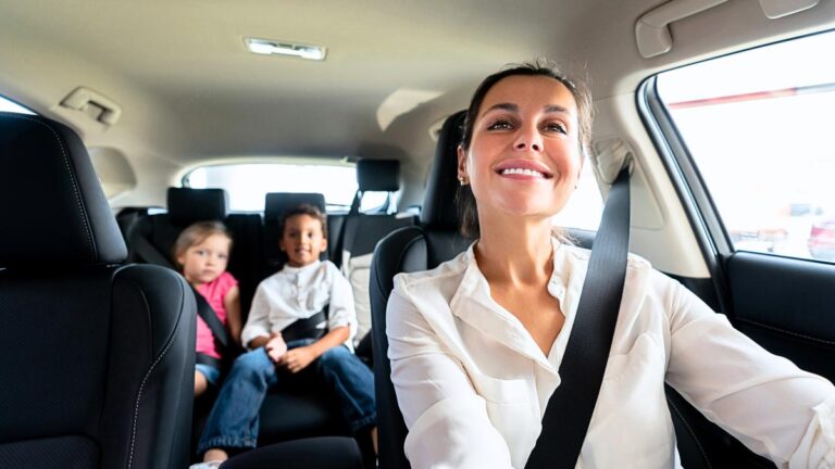 Smiling mom driving car with two kids buckled in the back seat.