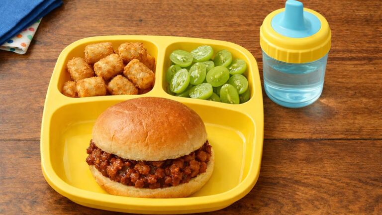 Kid-friendly lunch with a sloppy joe sandwich, tater tots, green grapes, and a sippy cup on a yellow tray.