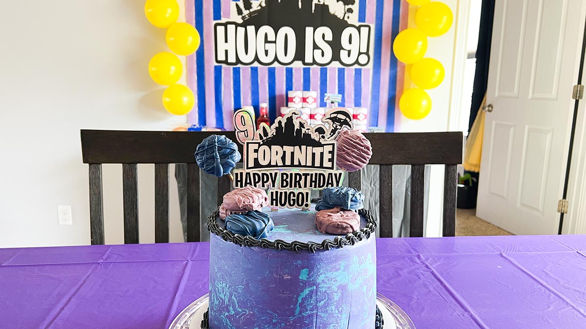Fortnite birthday cake on table with Hugo is 9 backdrop and balloons