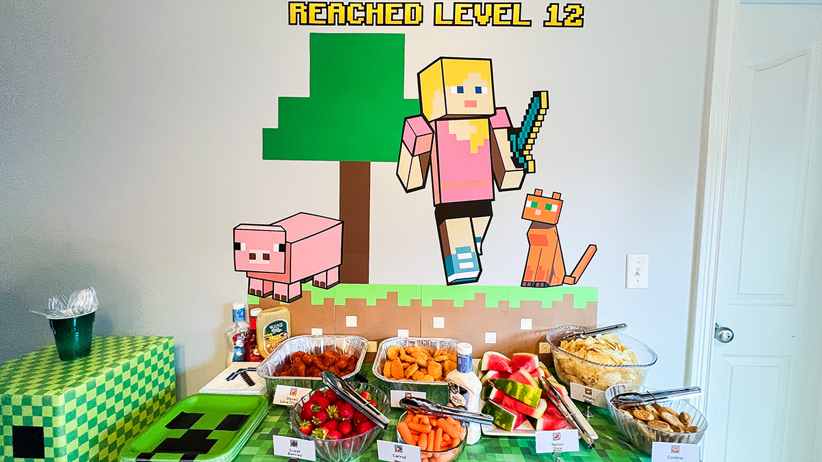 Minecraft birthday food table beneath a pixel backdrop with themed chicken, fruit, vegetables, chips, and cookies.