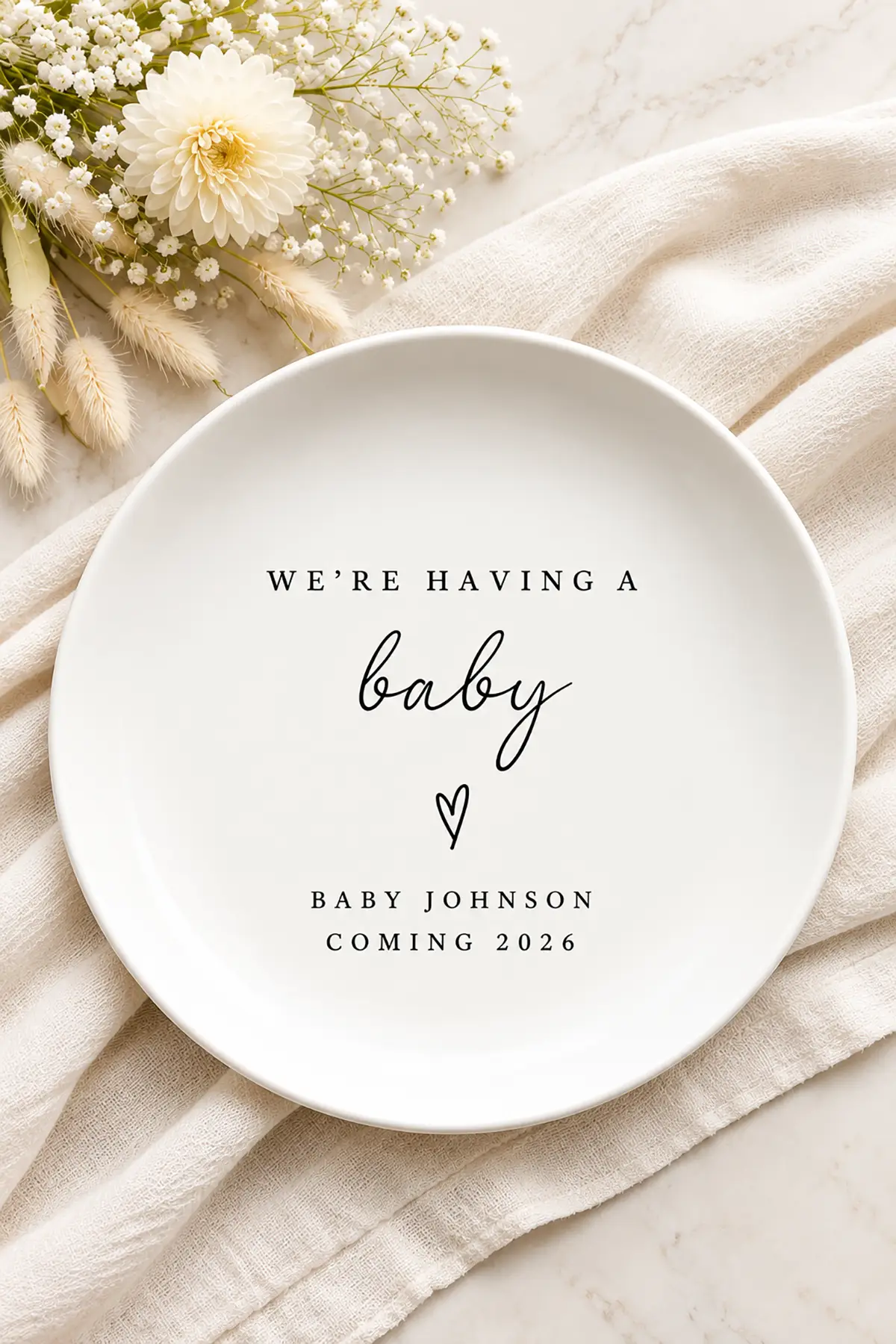 White pregnancy announcement plate reading "We're Having a Baby" and "Baby Johnson Coming 2026" with flowers and cream fabric.