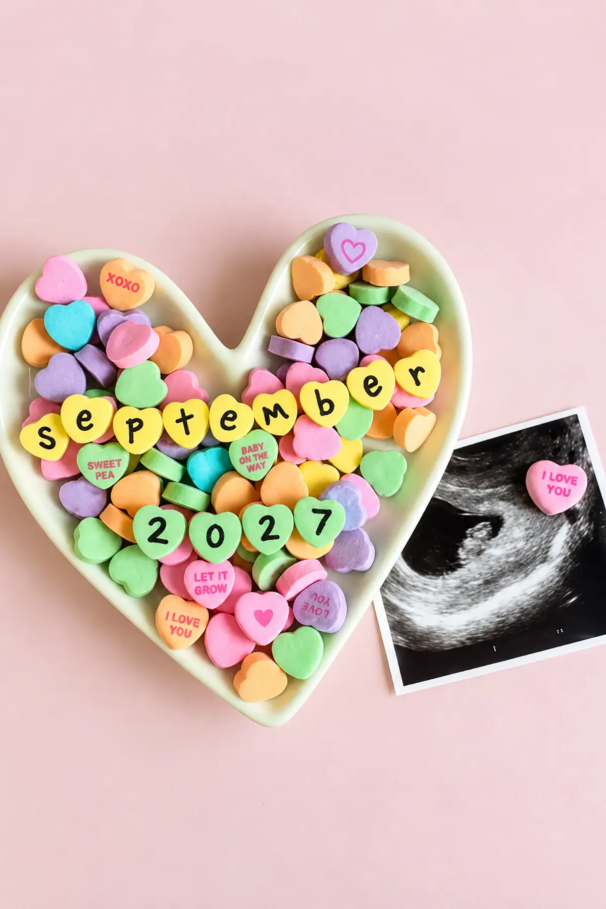 Colorful conversation hearts spelling "September 2027" in a heart-shaped dish beside an ultrasound for a pregnancy reveal.