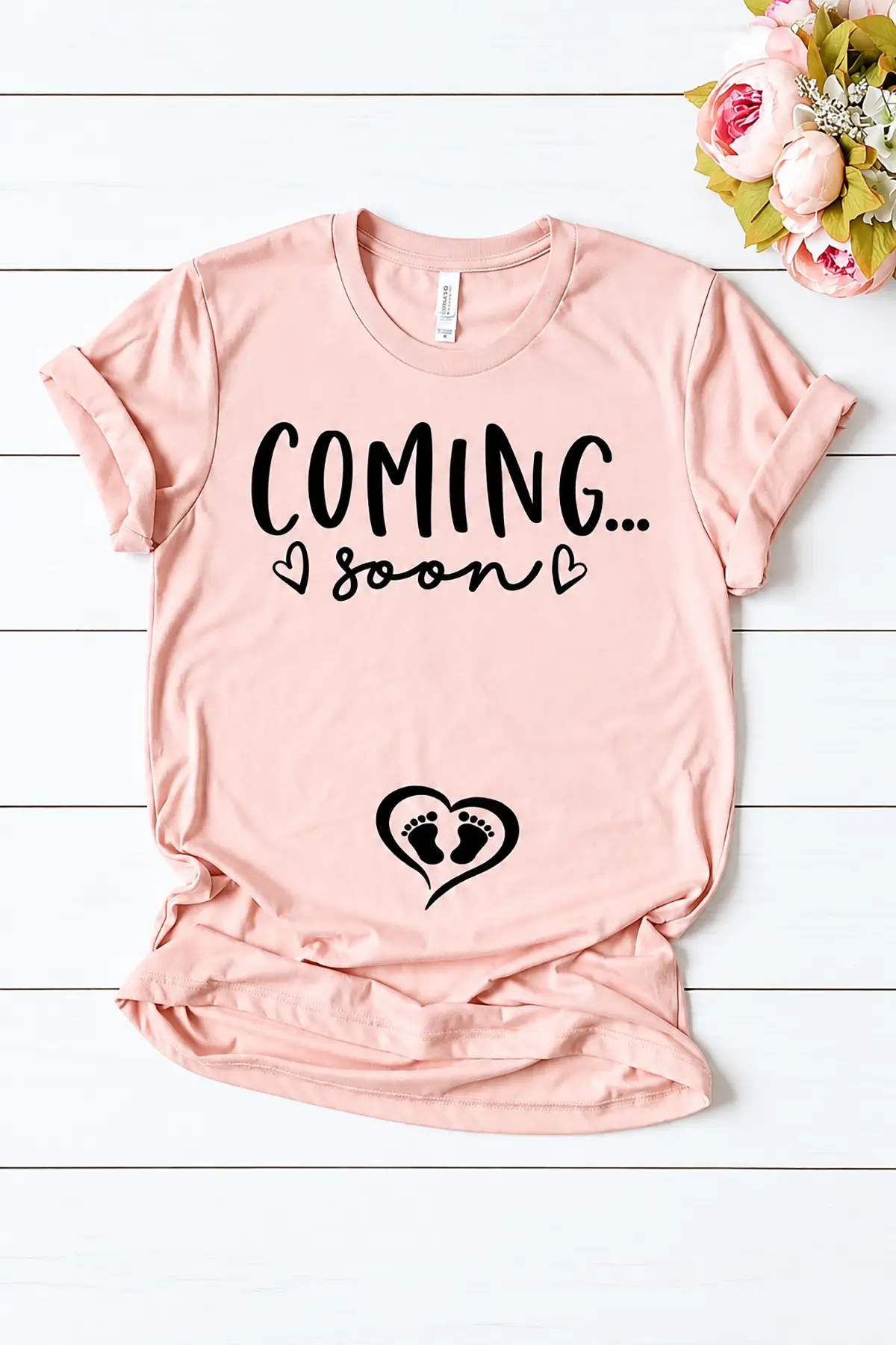 Pink "Coming Soon" pregnancy announcement T-shirt with baby footprints and heart design, displayed with flowers on white wooden planks.