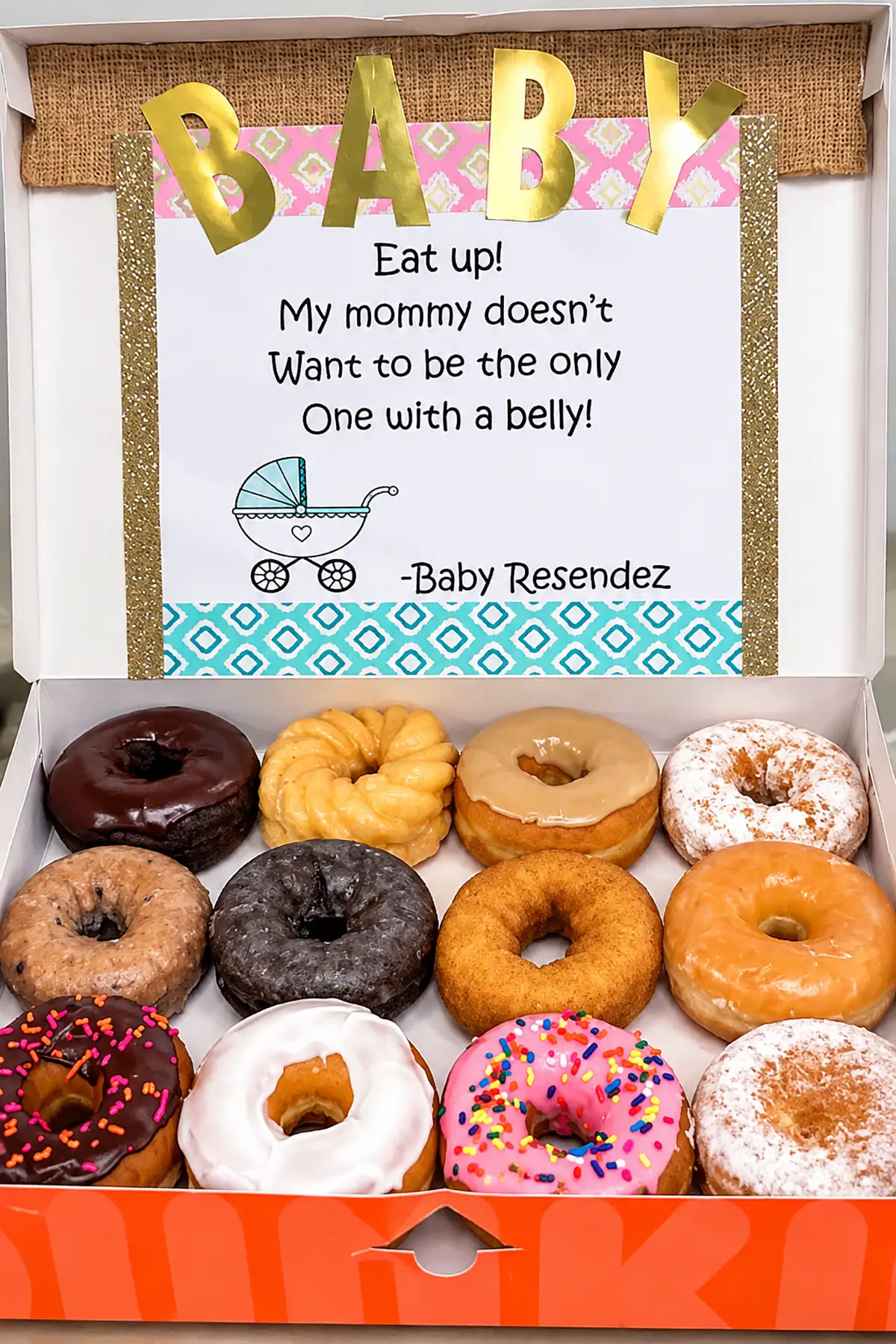 Open box of assorted donuts with a "BABY" pregnancy announcement sign telling Dad he won't be the only one with a belly.